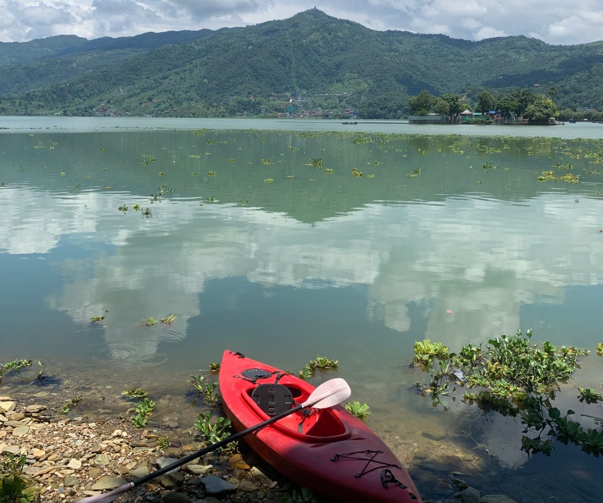 Red kayak resting on the rocky shore of Phewa Lake in Pokhara, Nepal with green hills and clouds reflected in the water