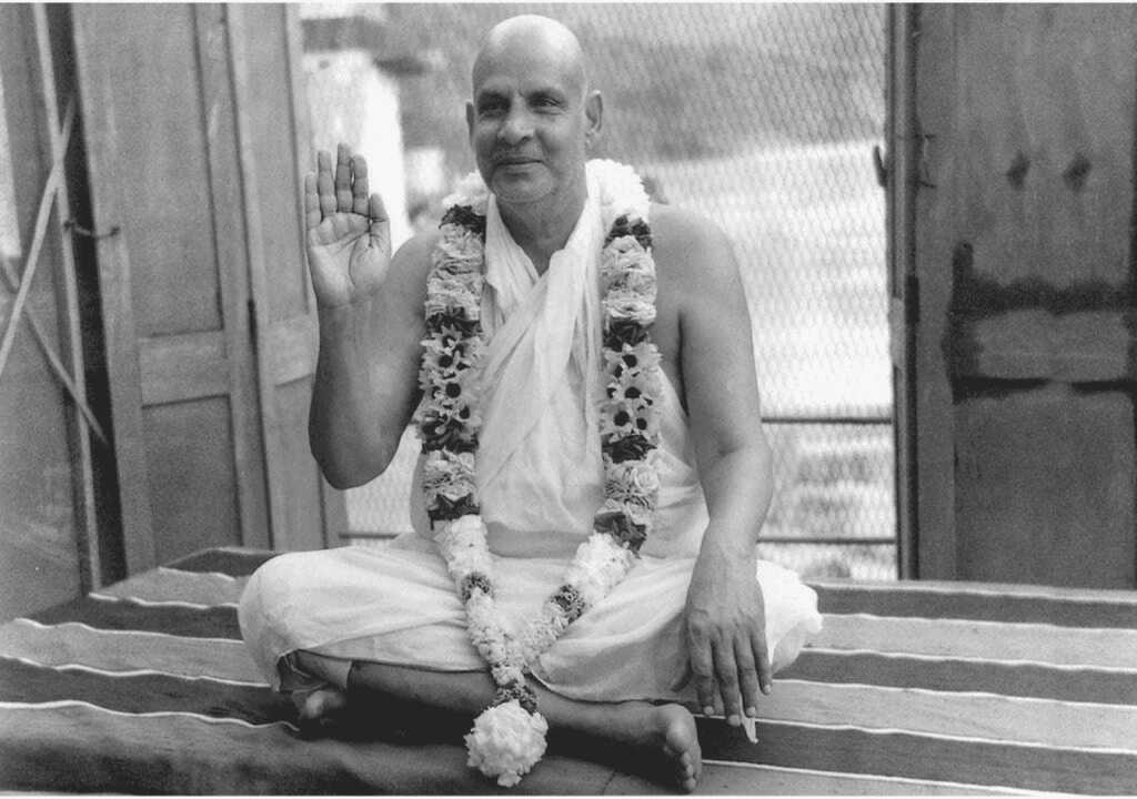 Swami Sivananda seated in a meditative pose with garlands, offering a raised hand in blessing