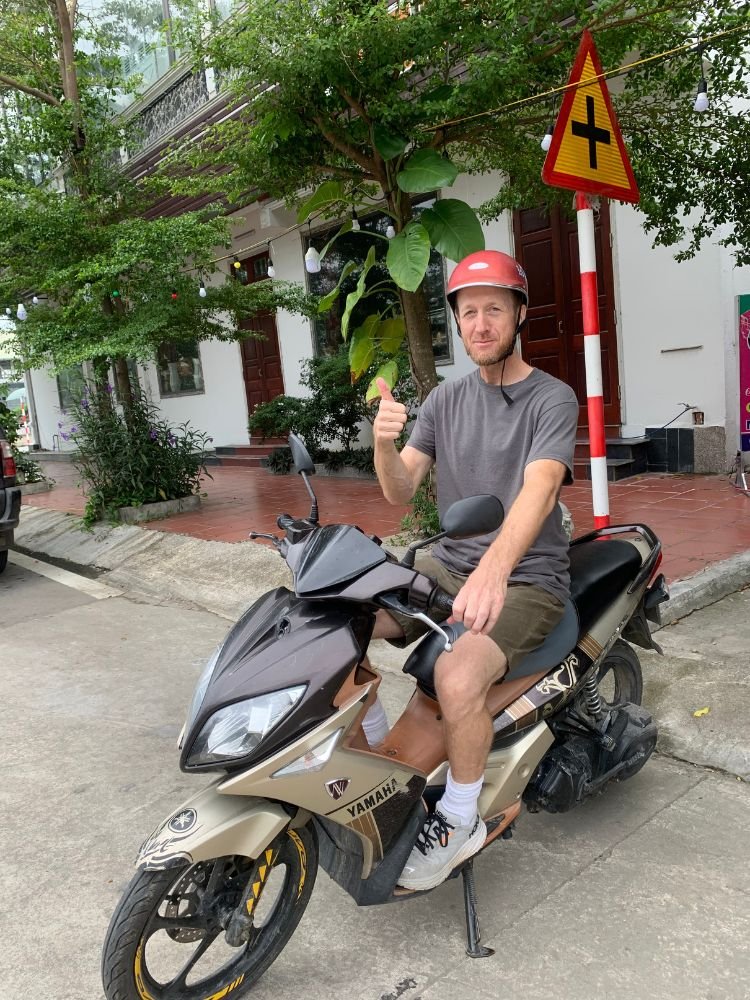 Traveler giving a thumbs up while riding a Yamaha scooter on a side street in Vietnam.