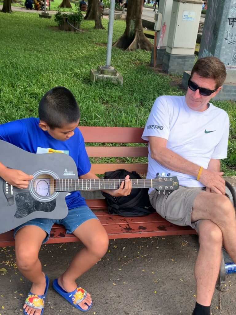Young Vietnamese boy plays a black acoustic guitar on a park bench beside a foreign man in sunglasses