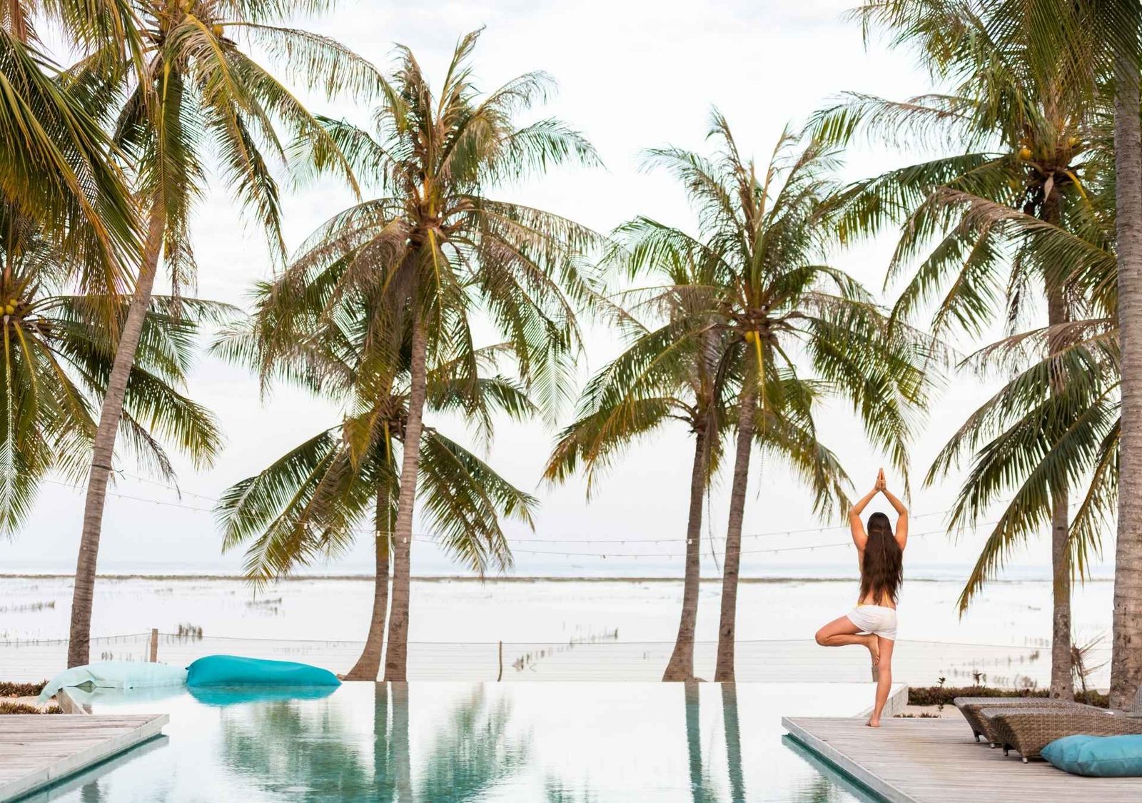 Woman practicing yoga beside an infinity pool under palm trees at a beachfront retreat