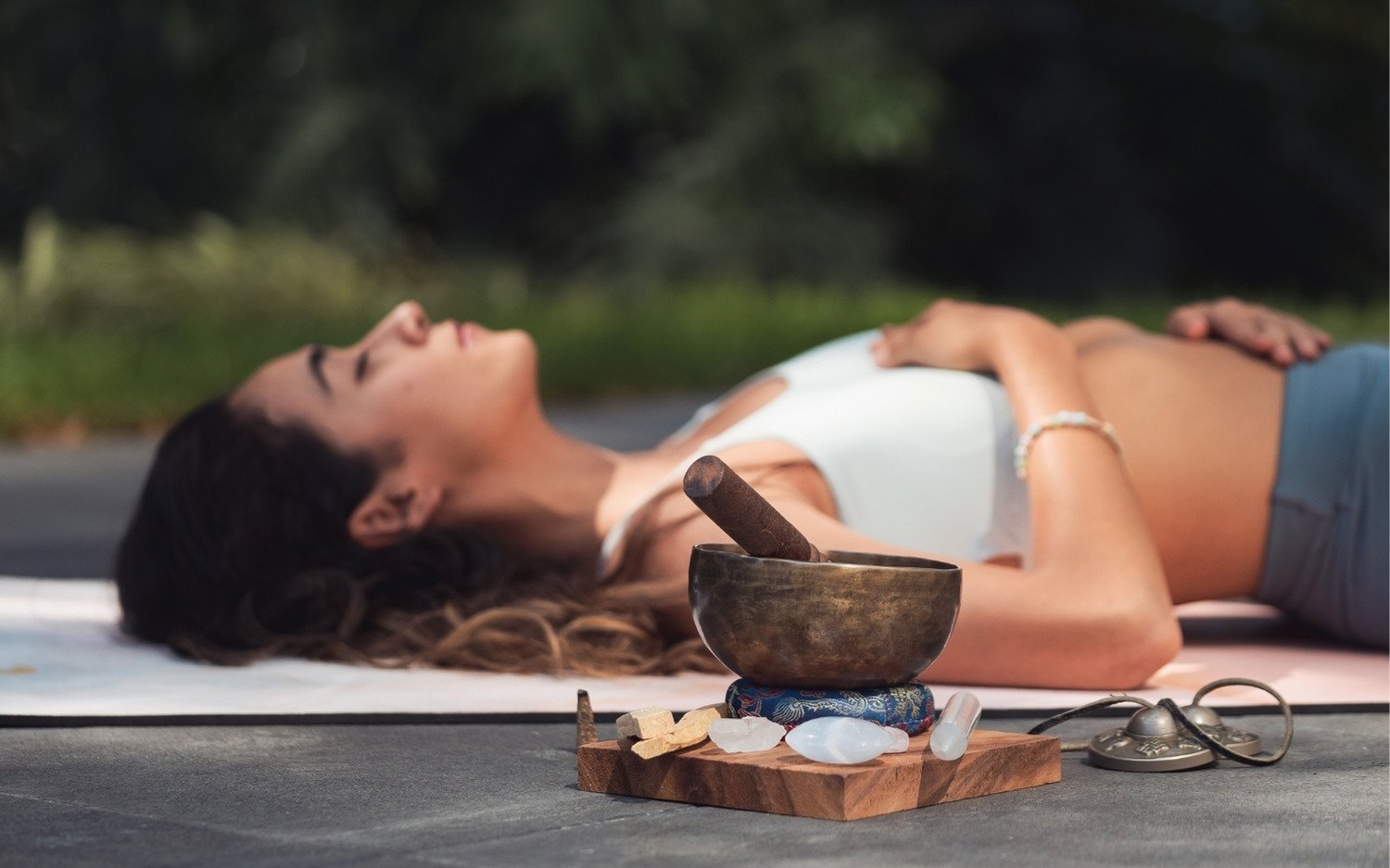 ytt-self-care-savasana-sound-bowl Yoga trainee lying in savasana outdoors with a sound bowl in the foreground
