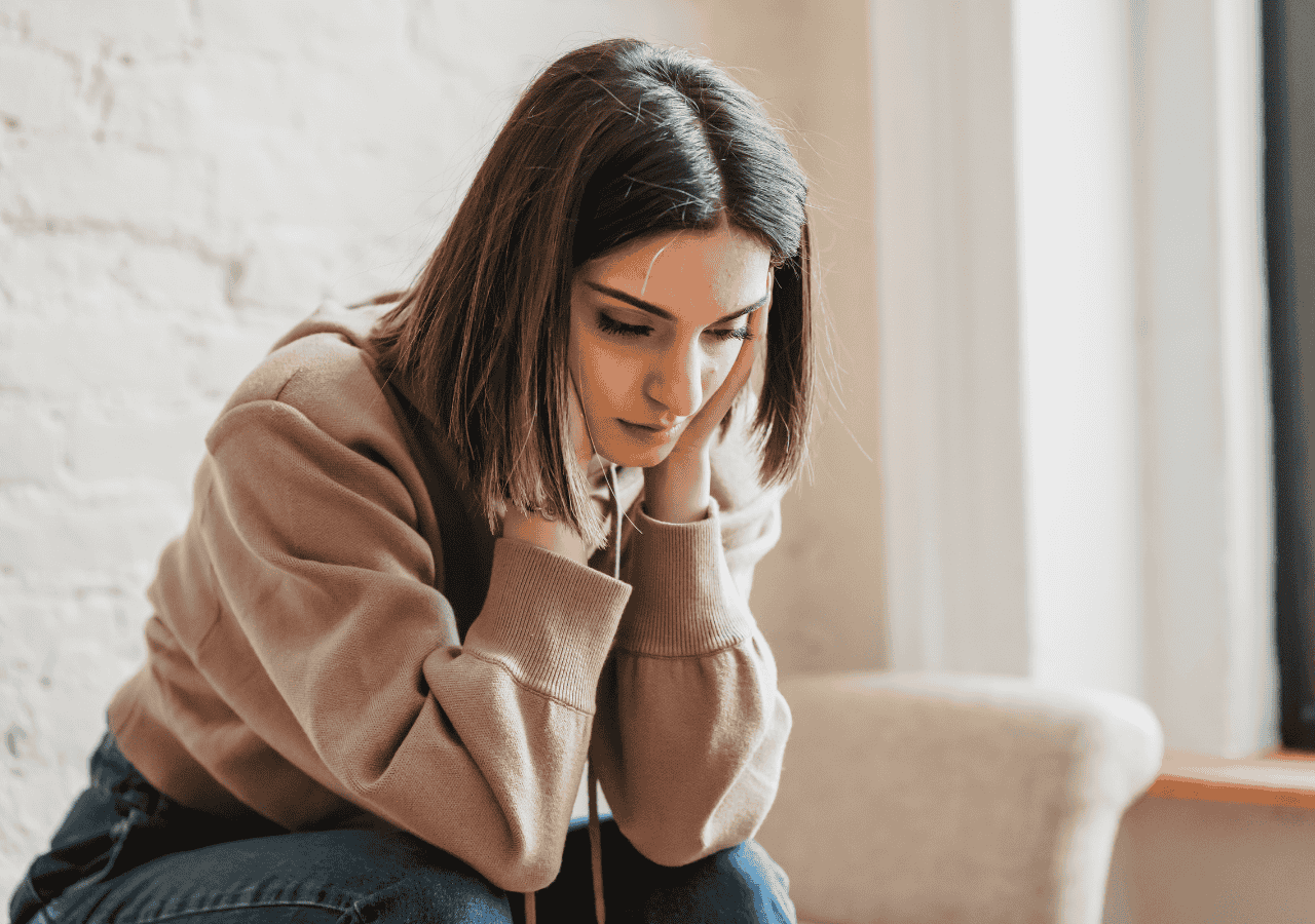 Woman sitting indoors looking down with a thoughtful, disappointed expression.
