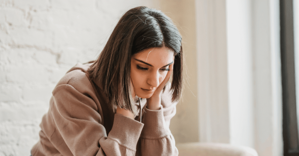 Woman sitting indoors looking down with a thoughtful, disappointed expression.
