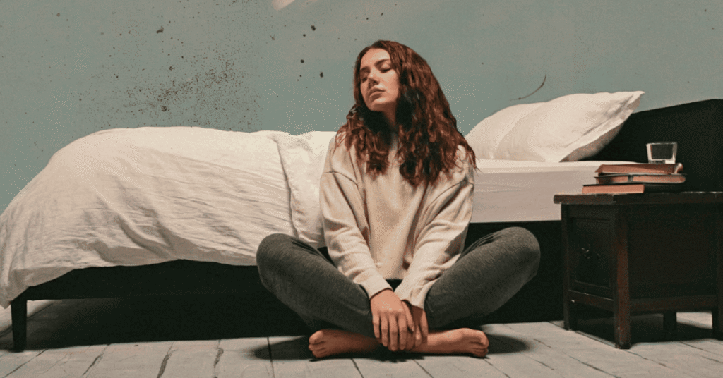 Woman sitting cross-legged on the floor by her bed with eyes closed in morning light