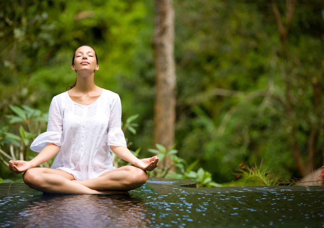 Woman meditating outdoors in white clothing by water and greenery