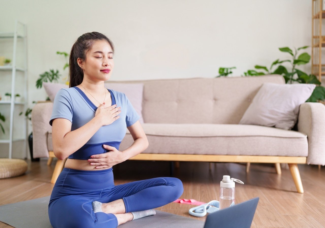 woman sitting cross-legged with one hand on chest and one on abdomen practicing deep breathing indoors